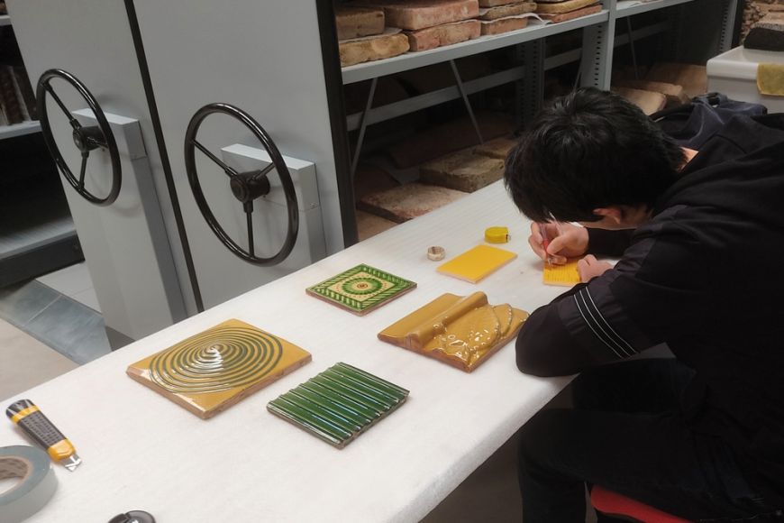 Inventariando piezas de cerámica de revestimiento de la colección Jaume Vilà en el fondo del Terracotta Museu | © Terracotta Museu Mesa con piezas de revestimiento y hombre de espaldas llenando fichas de inventario